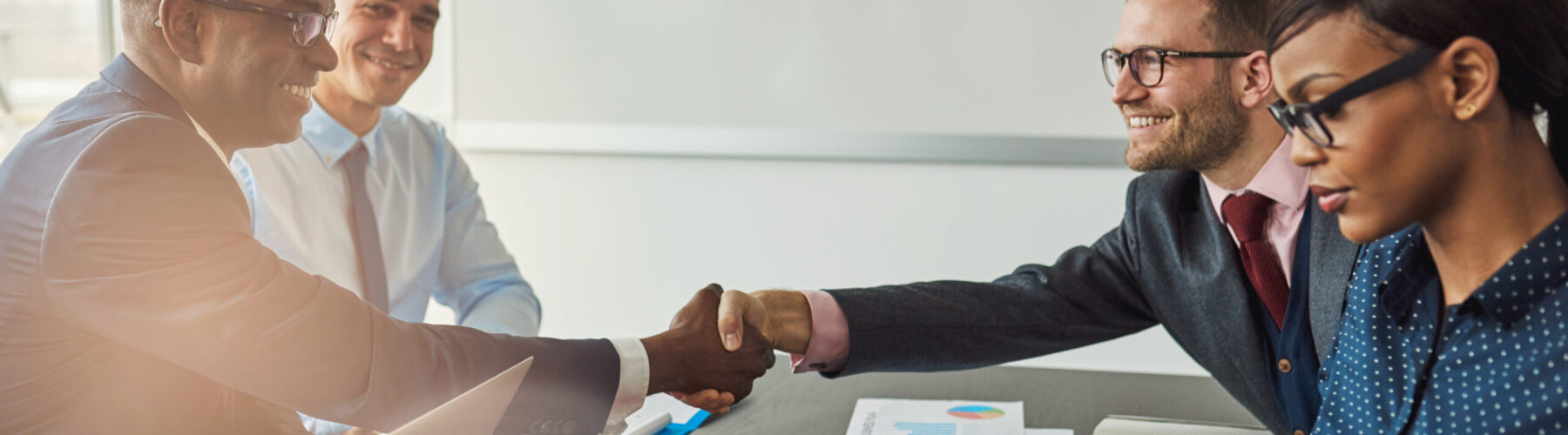 Multiracial team concluding a business agreement reaching across the table to shake hands with pleased smiles with focus to a young African woman in the foreground reading a tablet-pc